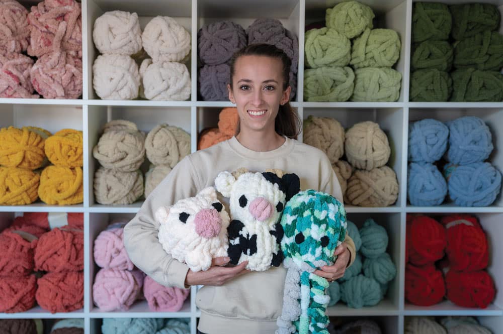 Sabrina stands in front of a wall of yarn with three of her handmade stuffies in her arms