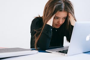 woman wearing a black shirt in a white room appears stressed while staring at a laptop screen.