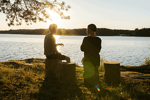 two people sitting next to a lake at sunset and talking